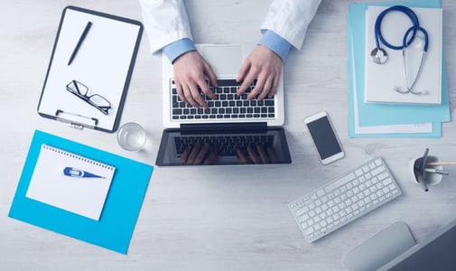 Dokter-met-medische-spullen-op-bureau-werkend-op-laptop-Unsplash (1)